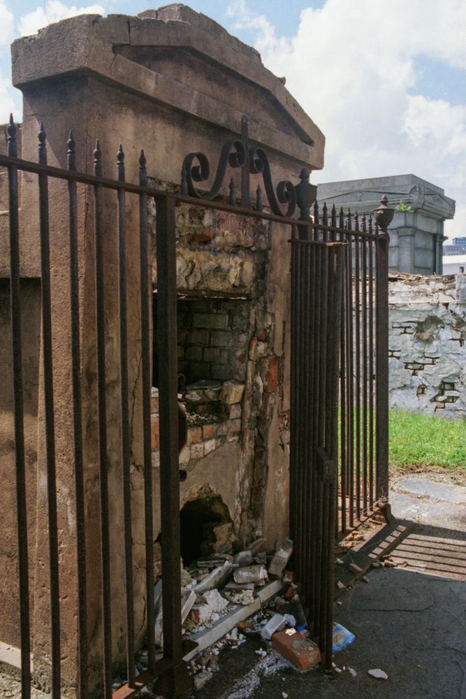 A three-quarters view of the front of a reddish two vault plastered brick tomb. A tall gated iron fence, rails topped with spikes, surrounds the tomb. The gate is partly opened. Both vaults have been smashed open, with rubble and brick on the ground in front. The open vaults disappear into darkness.