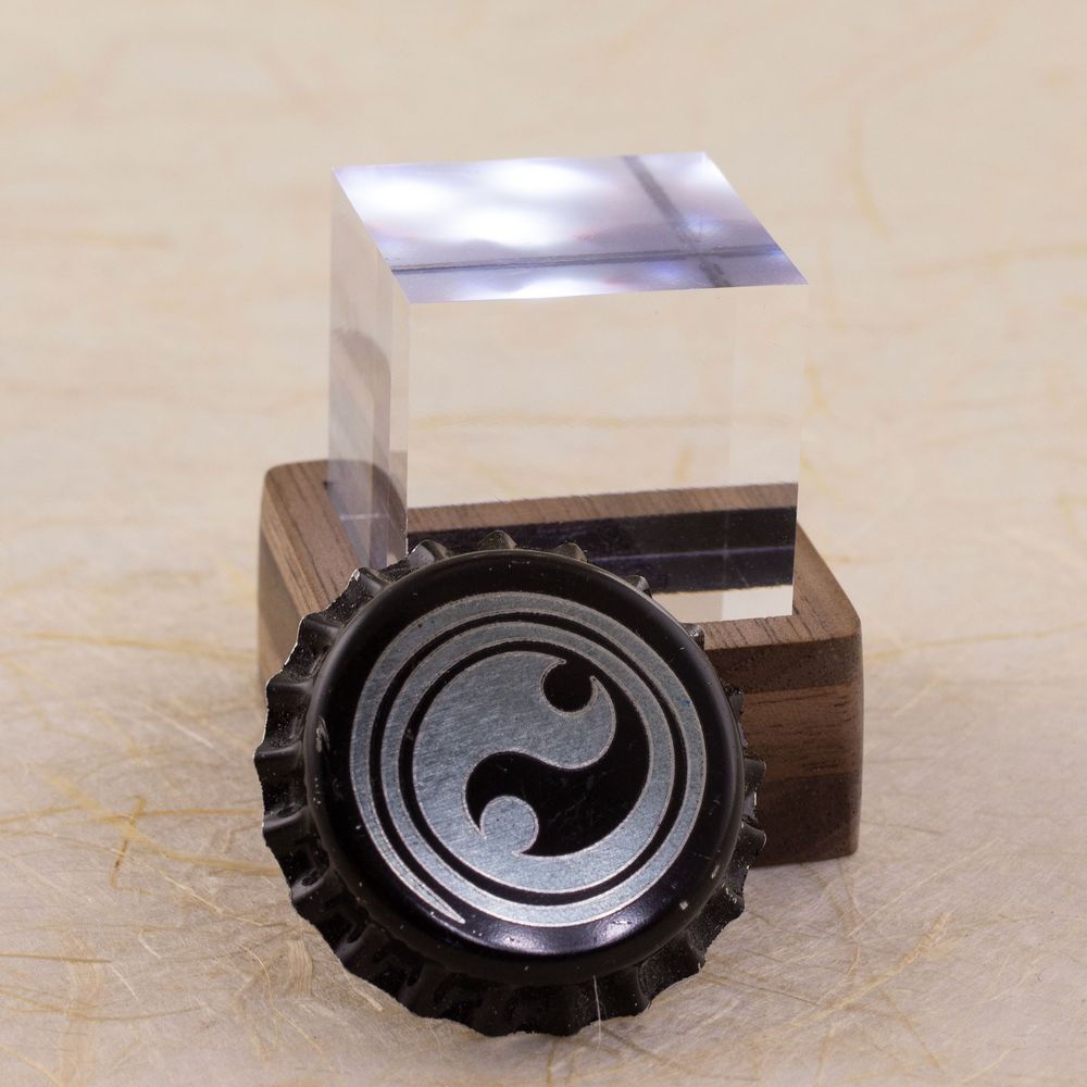 Side view of a one-inch clear acrylic cube sitting in a small square layered walnut stand. The stand lights the cube from below. There is a bottle cap with a Celtic spiral design engraved on it leaning against the stand to show scale. Shown against a defocused background of handmade paper with fiber inclusions. Side view.
