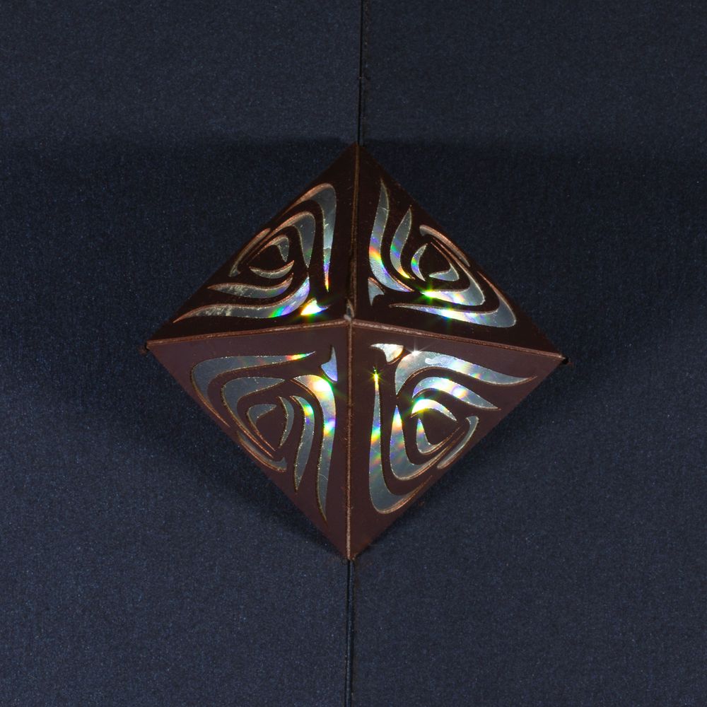 A tall, thin metallic copper four-sided pyramid with curvy organic cut outs on each face sits on a slightly pearlescent dark blue surface. The pyramid is lit from the inside, visible through the cut outs, with the light diffracted into rainbow shards. Top-down view from directly overhead showing all four faces of the pyramid.