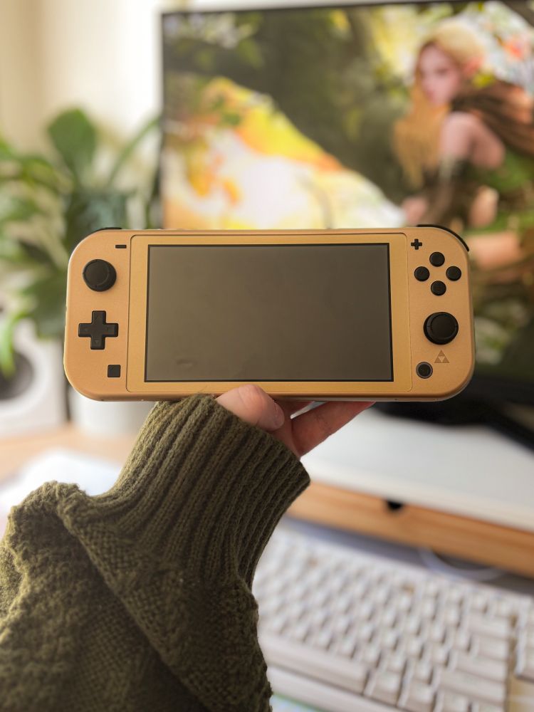 a photo of jade holding the nintendo switch lite hyrule edition in the foreground, with her desk setup unfocused in the background.