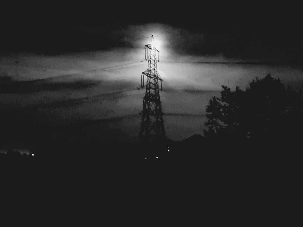 Cloud-hidden Full moon shining through the crown of a UK electricity pylon, using greyscale filter