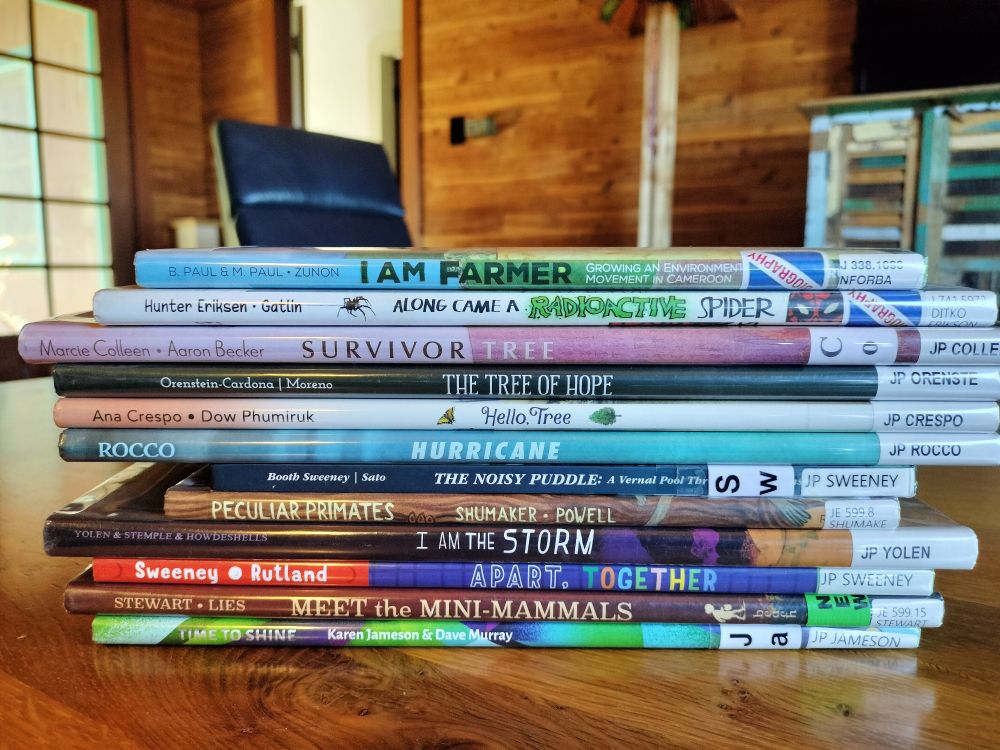A stack of pictures books on a table, with a room behind. Titles: I AM FARMER; ALONG CAME A RADIOACTIVE SPIDER; SURVIVOR TREE; THE TREE OF HOPE; HELLO, TREE; HURRICANE; THE NOISY PUDDLE; PECULIAR PRIMATES; I AM THE STORM; APART, TOGETHER; MEET THE MINI ANIMALS; TIME TO SHINE. 