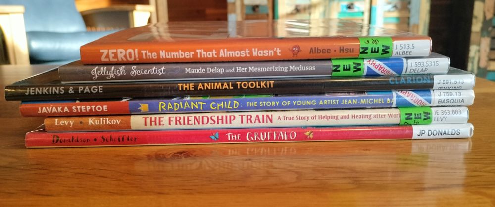 A stack of six library books on a table: ZERO: THE NUMBER THAT ALMOST WASN'T; JELLYFISH SCIENTIST: MAUDE DELAP AND HER MESMERIZING MEDUSAS; THE ANIMAL TOOLKIT; RADIANT CHILD: THE STORY OF YOUNG ARTIST JEAN-MICHEL BASQUIAT; THE FRIENDSHIP TRAIN: A TRUE STORY OF HELPING AND HEALING AFTER WORLD WAR II; THE GRUFFALO