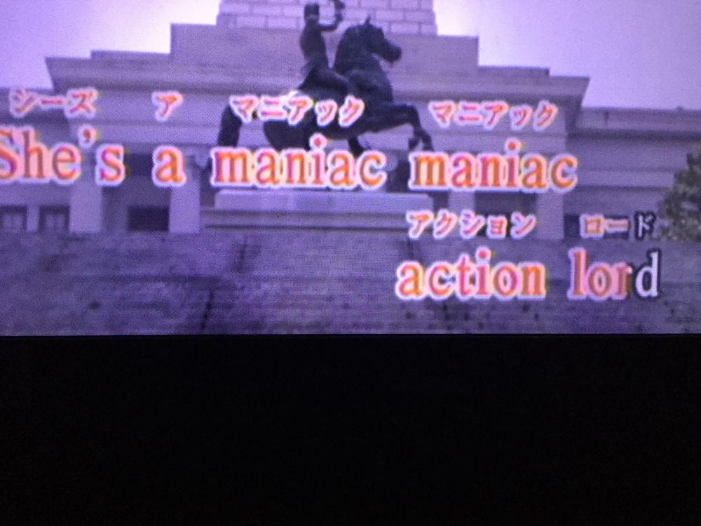 photo of a karaoke screen, on the chorus of Maniac by Michael Sembello. there is katakana ruby above the English words and footage of a statue in the background. instead of the correct lyrics “she’s a maniac, maniac at your door” it reads “she’s a maniac, maniac action lord”