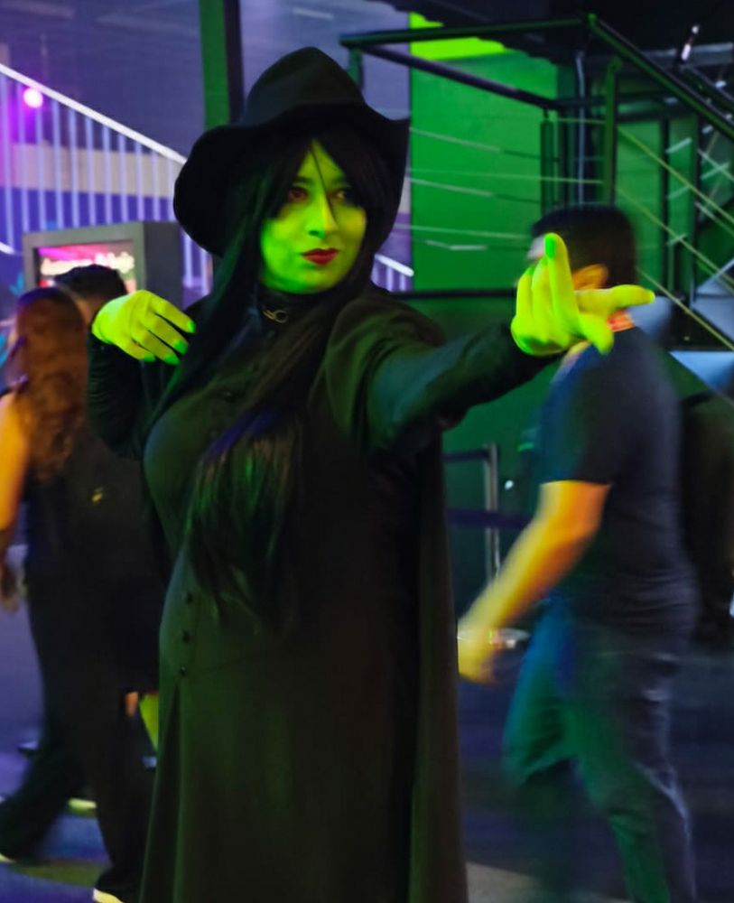 Uma foto minha vestida de Elphaba, do musical Wicked. Estou com vestido longo preto de mangas compridas, capa preta, chapéu de bruxa pontudo, cabelos pretos compridos e pele verde.
