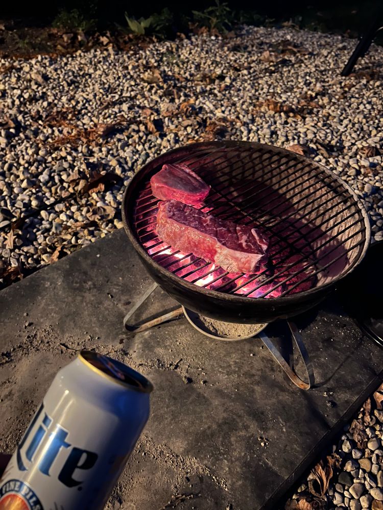 Steaks on a mini grill in the dark with a can of Miller Lite in the foreground 
