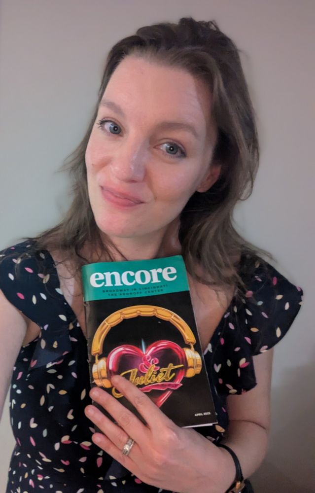 A woman is smirking and holding a "playbill" for &Juliet