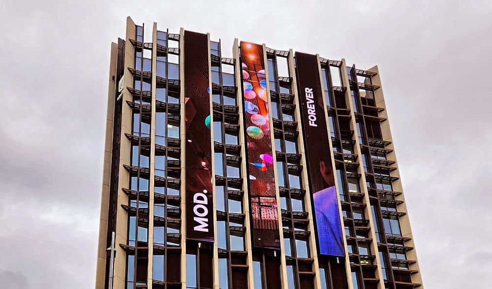 Viewing the Museum of Discovery from below, a tall concrete, steel, and glass building with its abbreviated name in a multi colored light display 