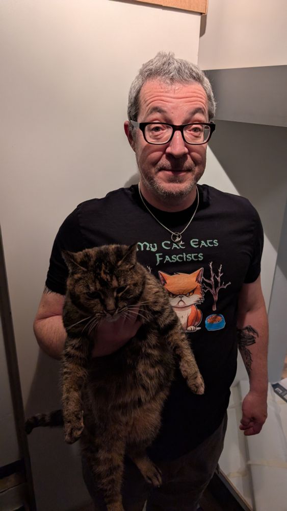 A picture of me a white presenting middle aged male with gray hair and black glasses wearing a black shirt that says my cat eats fascists while I'm holding a tabby cat in my hand. She does. Oh so she very much does.