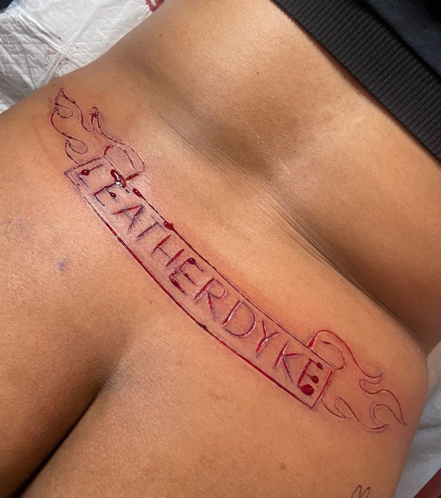 The lower back of a Black person. The word “LEATHERDYKE” has been carved into the skin with a banner surrounding it. The wound is actively bleeding 