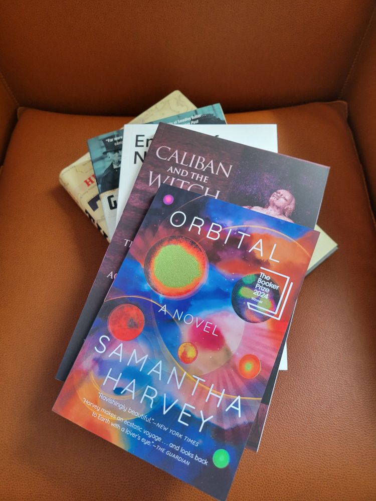 Five books in a slightly fanned out stack on an orange leather chair, the top book is Orbital by Samantha Harvey, with a cover of colorful planetary bodies in a blue space, the next book title is visible, Caliban and the Witch, with the picture of a woman's head visible, wearing an old fashioned bonnet, the rest of the book covers and titles are not visible.