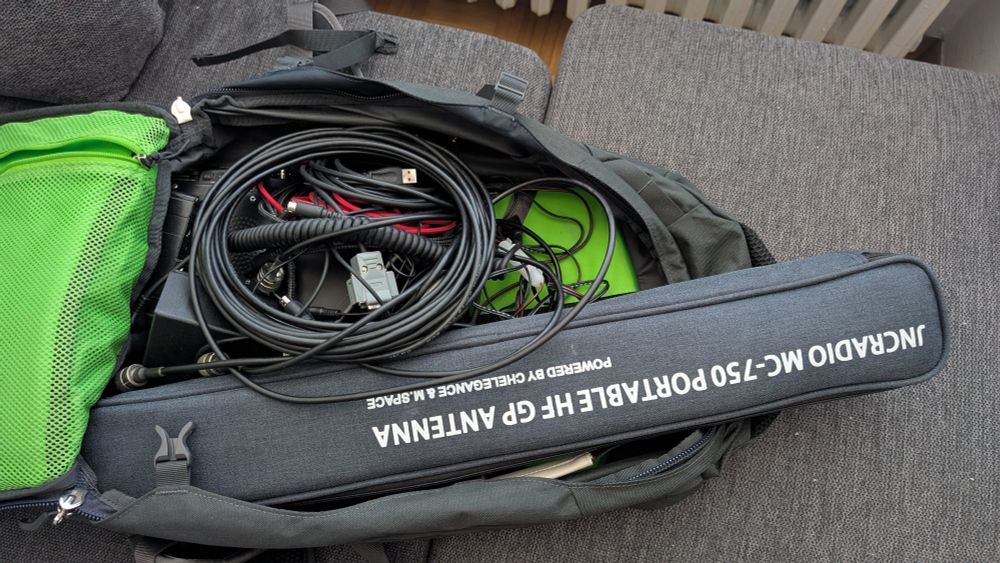 Foto eines offenen Rucksacks. Zu sehen sind viele überwiegend schwarze Kabel, darunter sind mehrere kleine schwarze Geräte zu erkennen. Obenauf liegt ein Futteral mit der Beschriftung "JNCRadio MC-750 Portable HF GP Antenna"