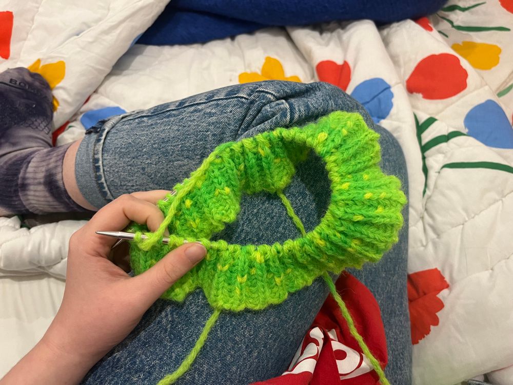 A hand holds the beginnings of a mottled acid green knit hat. The hand and hat rests on folded legs wearing blue jeans. The legs sit on white comforter decorated with brightly colored graphic flowers.