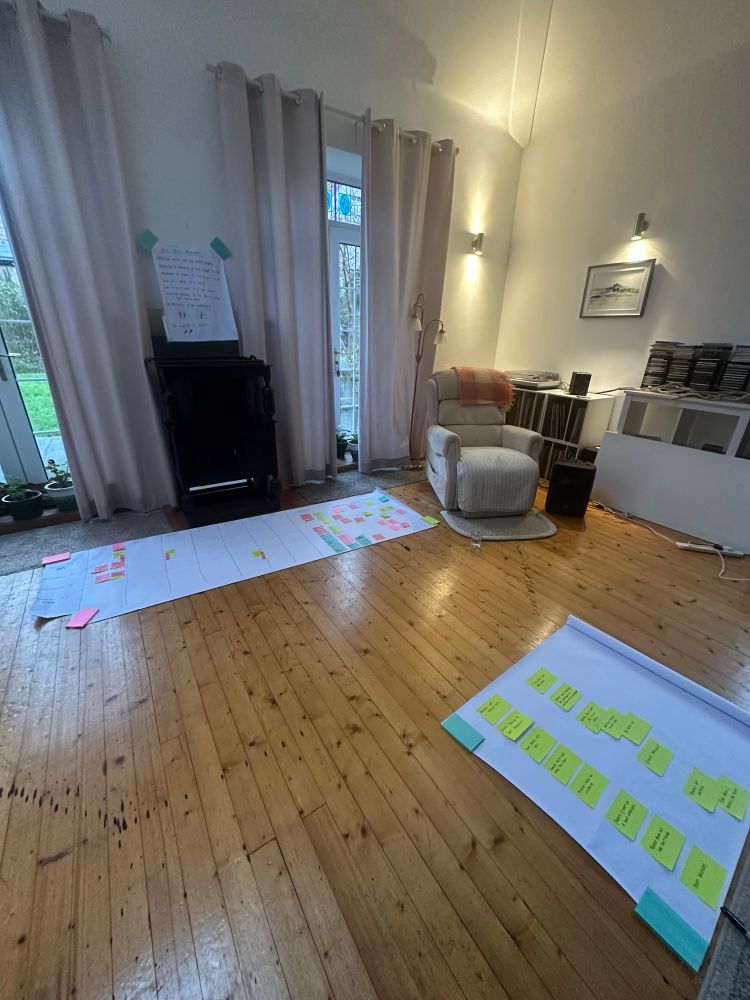 A high ceiling studio with wooden floorboards, and two windows. Soft lighting. There are large pieces of paper with post it notes on the floor. Another handwritten poster is stuck to a wall. 