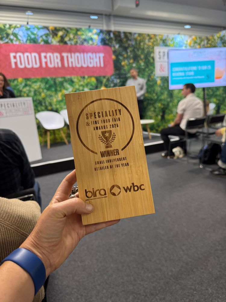 A hand holding the award, which is a block of wood, in front of a trade show stage, with words "Food for thought" on the wall.