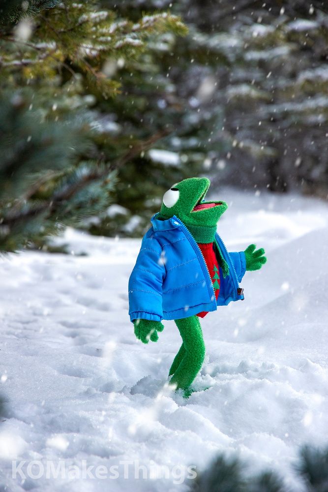 A replica of Robin the Frog, from the Muppets, standing in the snow surrounded by evergreen trees. Robin is looking up at the sky, watching falling snowflakes. Robin is wearing a blue puffy winter jacket, a red sweater with a green christmas tree on it, and a small green knit scarf. 