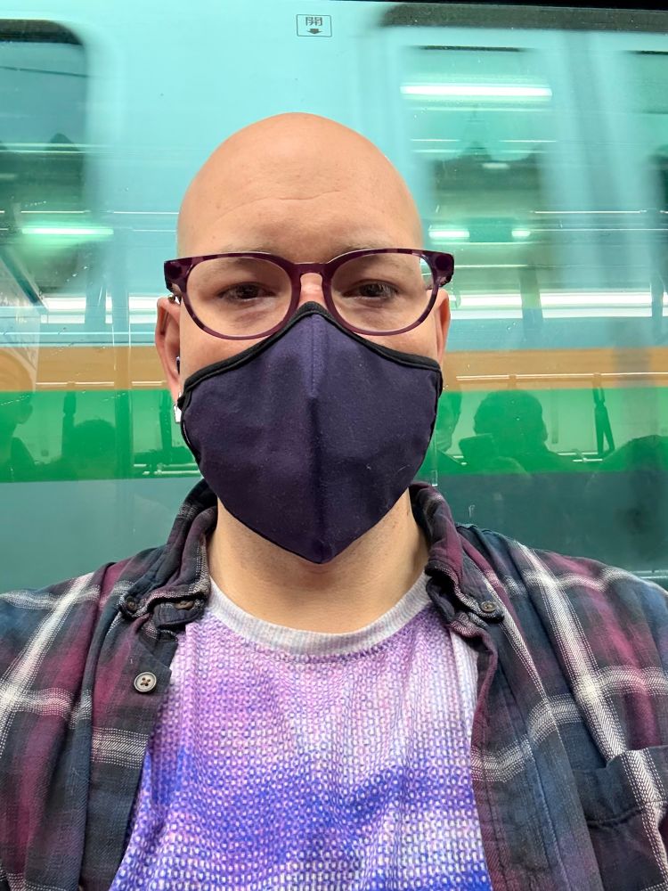 Train selfie. I’m wearing purple glasses, a navy mask, a “digital noise” print shirt with white, purple, and blue, and a blue, purple, and white flannel shirt. My head is shaved. 