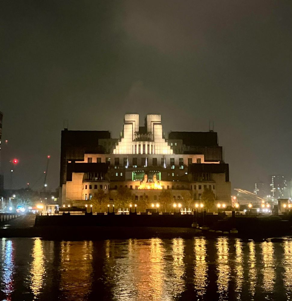 MI6 building as a dark silhouette with a few sections spotlit - seen from across the river with lights reflected in the water