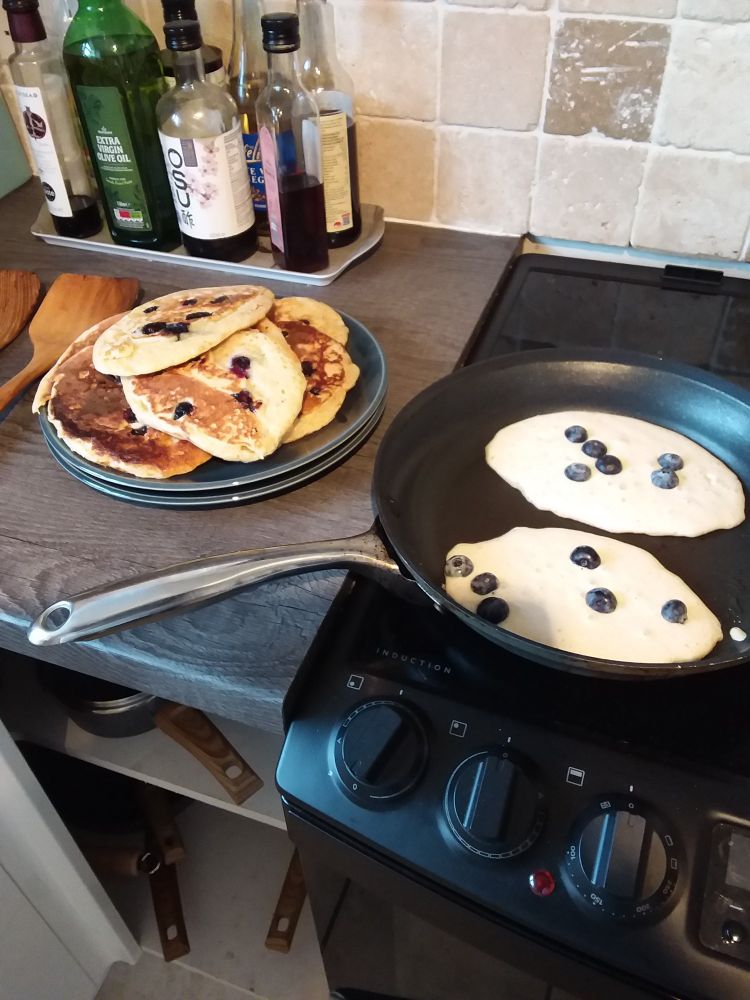 A pile of blueberry muffins on a plate and two frying in the pan.