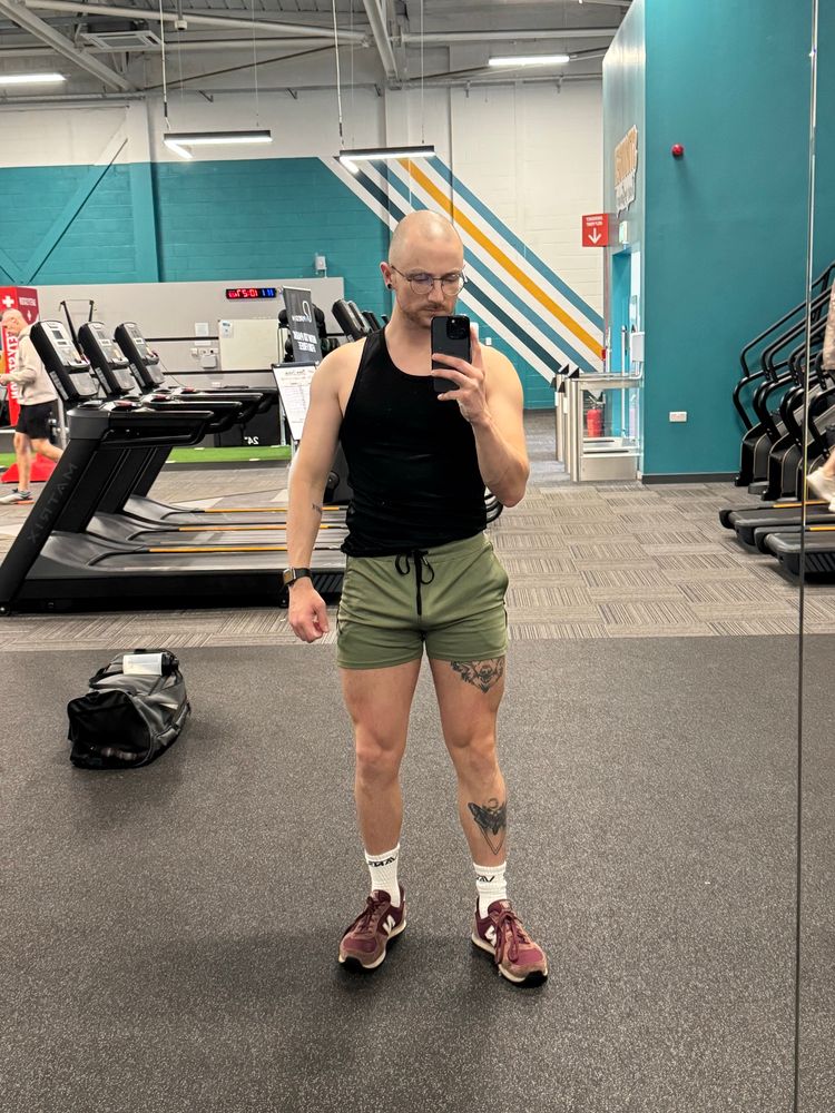 Gym selfie. I am stood in front of the gym mirror taking a picture with my phone in my right hand. I am wearing a black gym vest and khaki green shorts. I have white socks on with a pair of burgundy trainers on. In the background you can see a number of treadmills and stairmaster machines 