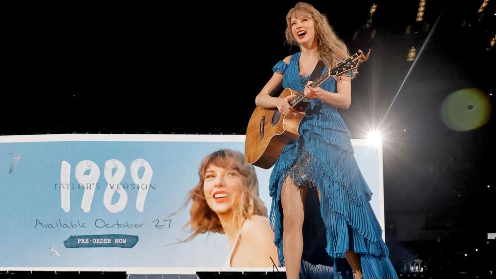 Taylor Swift on stage, holding a guitar and smiling. She’s wearing a blue dress.

Behind her on the screen is the announcement for 1989 Taylor’s version 