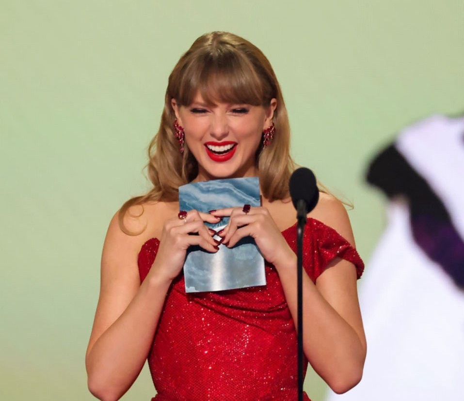 Taylor Swift smiling on stage at the 67th grammys, holding the envelope with the best country album winner