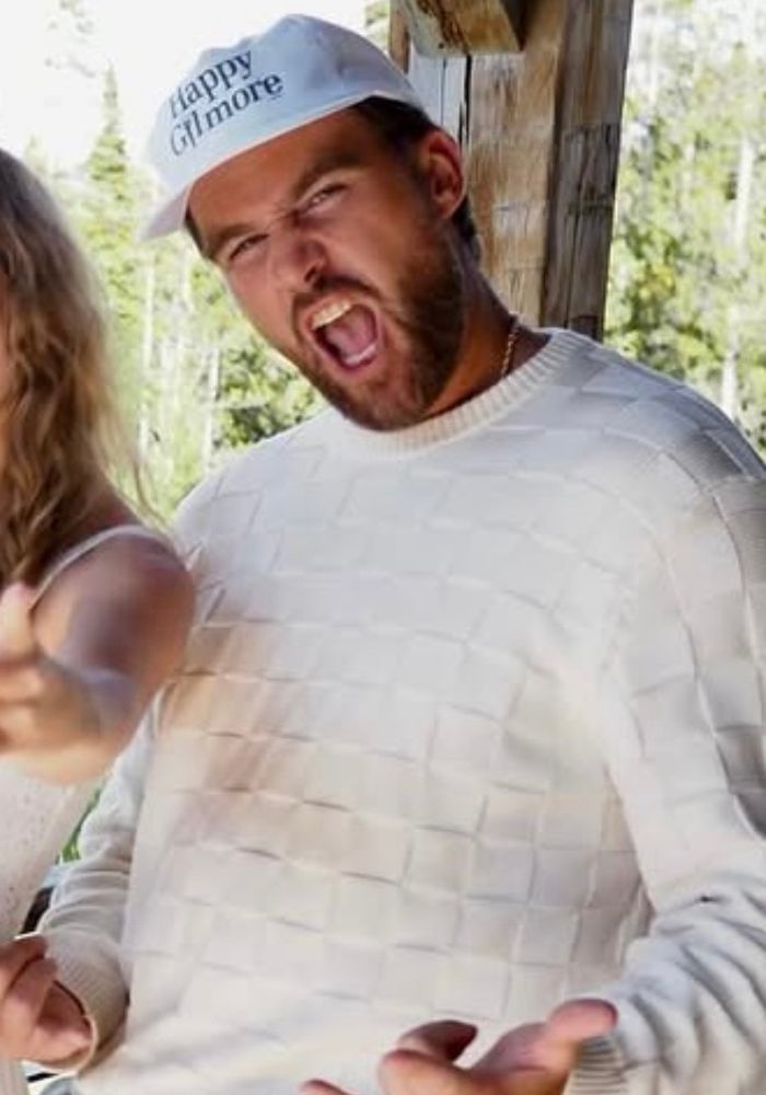 Travis Kelce in a white patterned sweater and "Happy Gilmore" cap is playfully making a shouting face and making a gesture with his hands, almost like he’s yelling hell yeah 