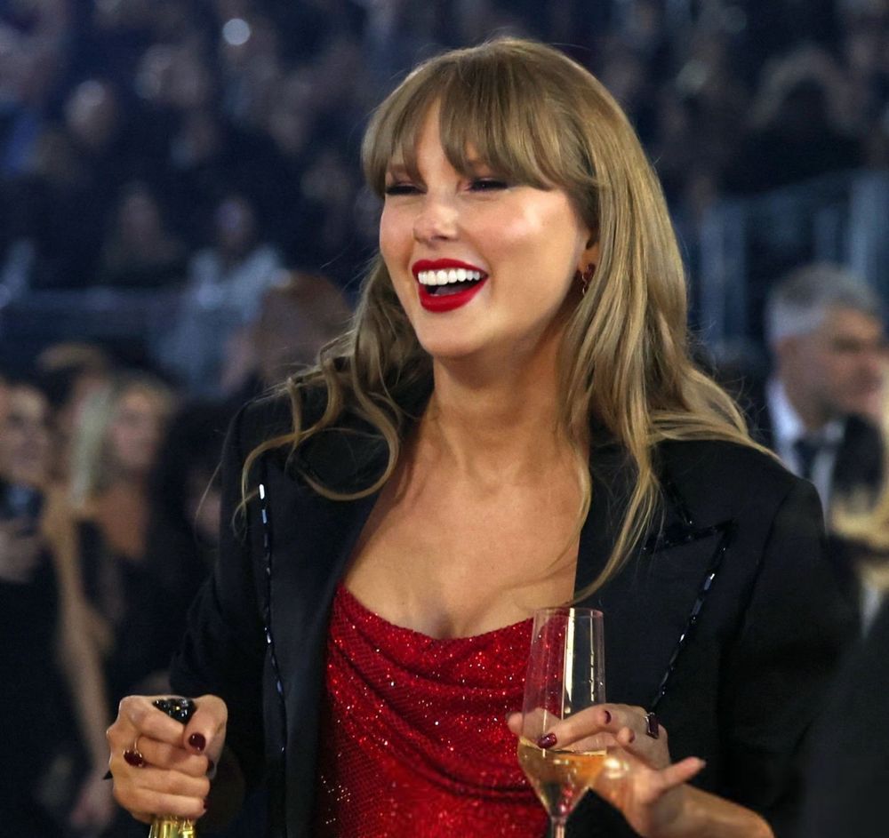 Taylor Swift smiling, holding a bottle and a glass of champagne with her pinkie finger sticking out 