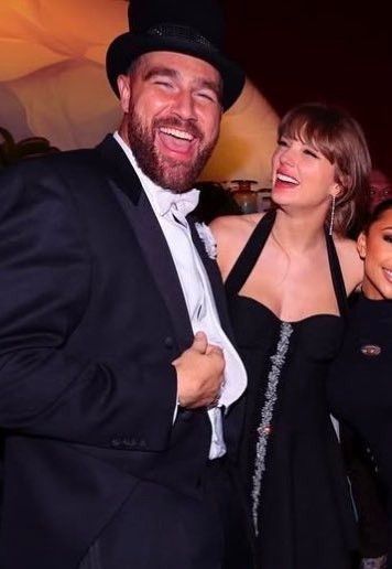 Picture of taylor swift and Travis Kelce. Travis is smiling big at the camera and taylor is looking at Travis
