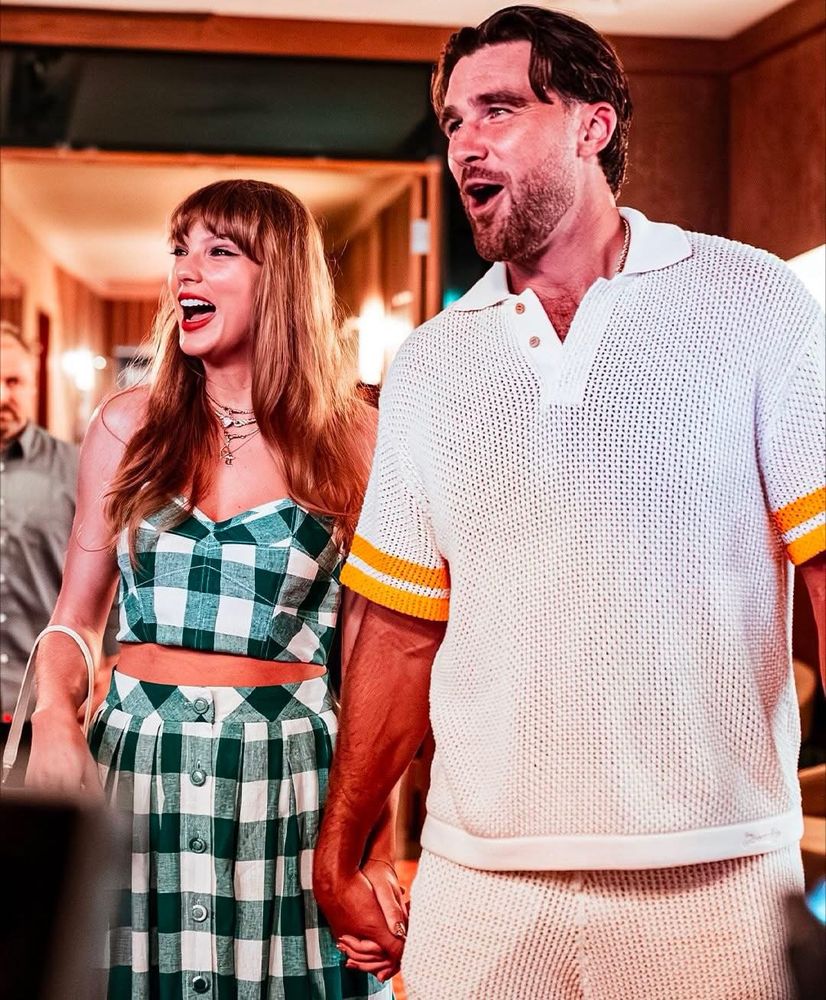 Taylor Swift and Travis Kelce holding hands and looking off camera making happy/wowed faces.

Taylor is in a green plaid dress and Travis is in a white shirt and cream coloured shorts 
