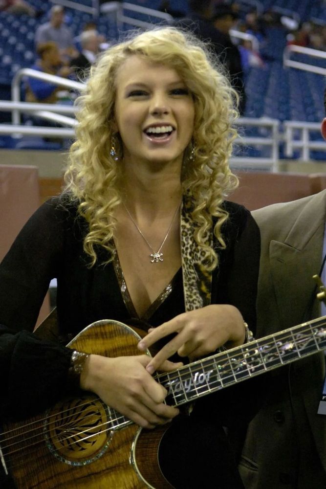 Picture of taylor swift from 2006-ish. Her hair is super curly and she’s wearing a black long-sleeve, holding a guitar in front of her and smiling