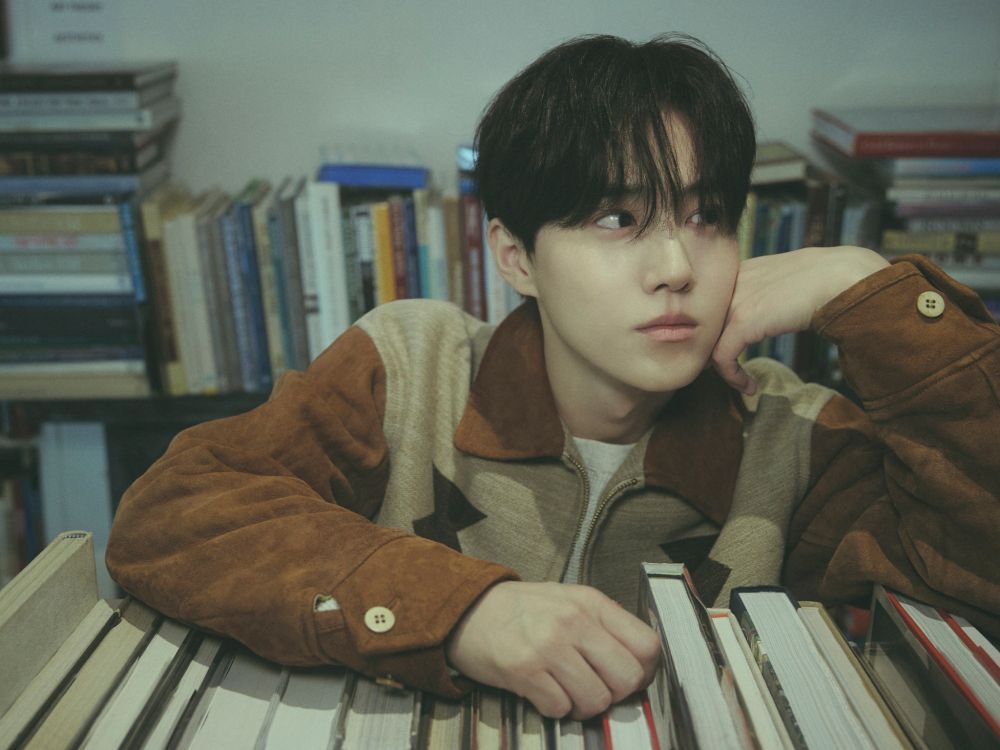 Suho of kpop group exo posing for a teaser image on top of books, where a brown suede jacket.