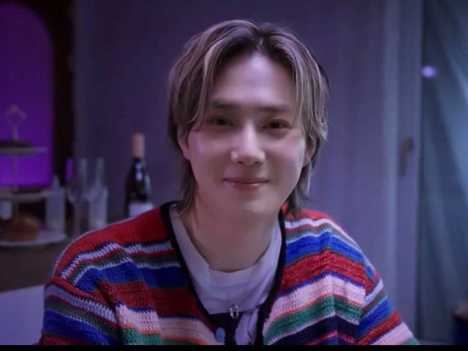 Portrait is Suho of EXO with blond highlights in his hair and a multicoloured striped cardigan. He'is lookout the camera with a soft smile.