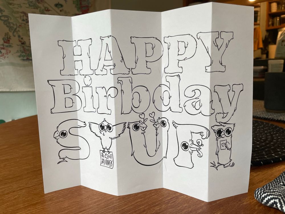 A folded paper printed with the words “Happy Birbday Stüfi” using letters made of feathers and birds. The word “birb” is cute internet slang for “bird”. The umlaut is made of heart flowers held by two birbs. 