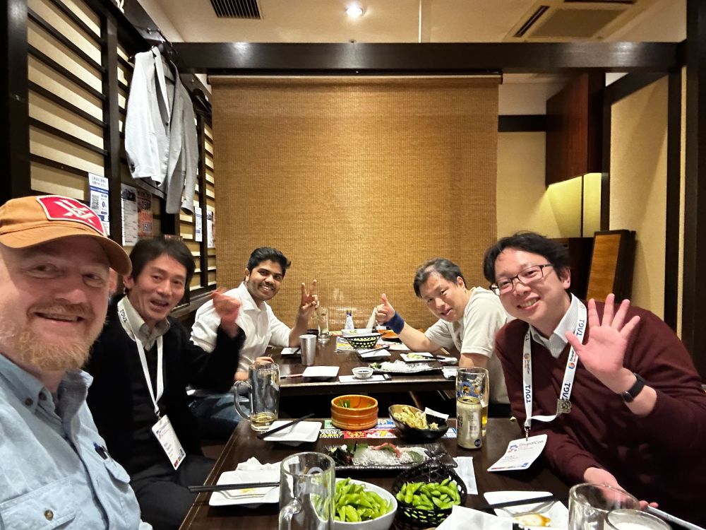 A group of DrupalCon attendees sit together in a cozy izakaya, smiling and waving at the camera. Plates of edamame, grilled skewers, and drinks cover the long wooden table. The atmosphere is warm and lively, capturing the camaraderie and comfort of a shared meal in Nara.