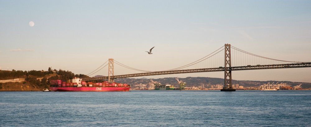 A small magenta cargo ship passes under the western span of the San Francisco Bay Bridge, in front of Yerba Buena Island. The sky is dusk and showing the Belt of Venus, the sea a gentle blue, a full moon is faint and low, as a gull flies through the frame above the bridge's suspension cables.