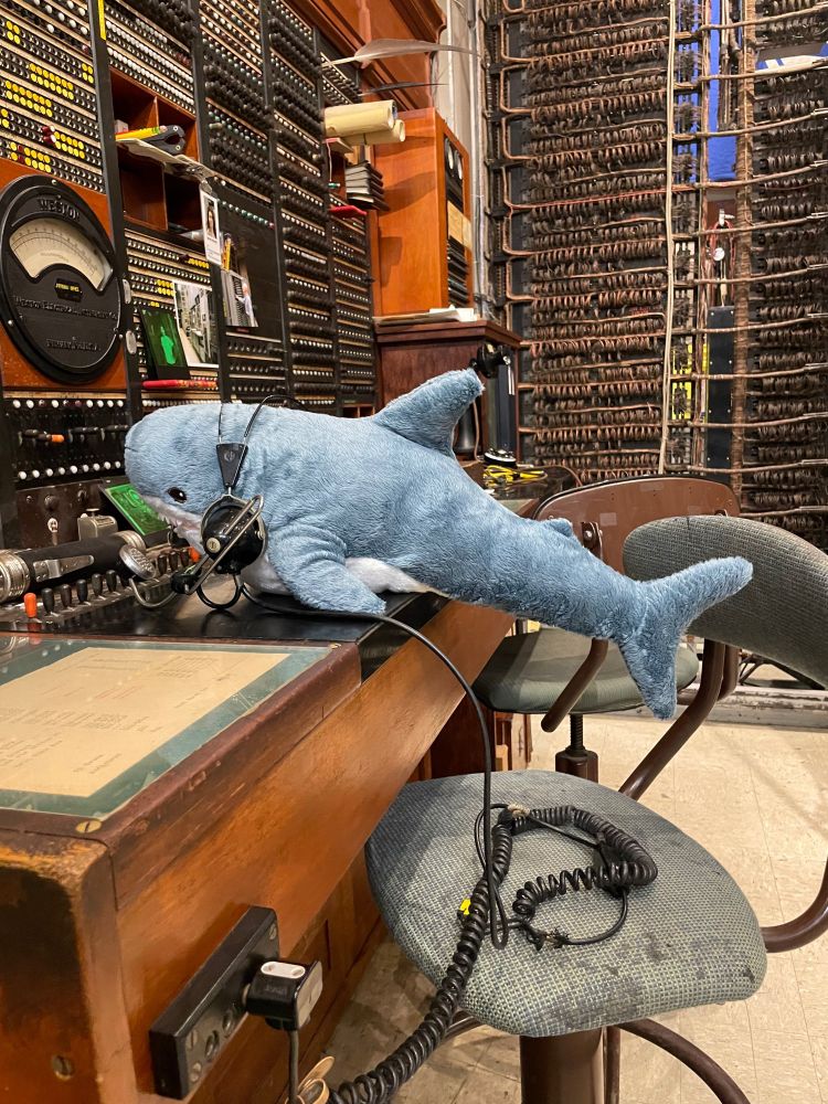 a small stuffed shark wears a headset and microphone at a wood panel technician’s desk for a telephone switch from 1923