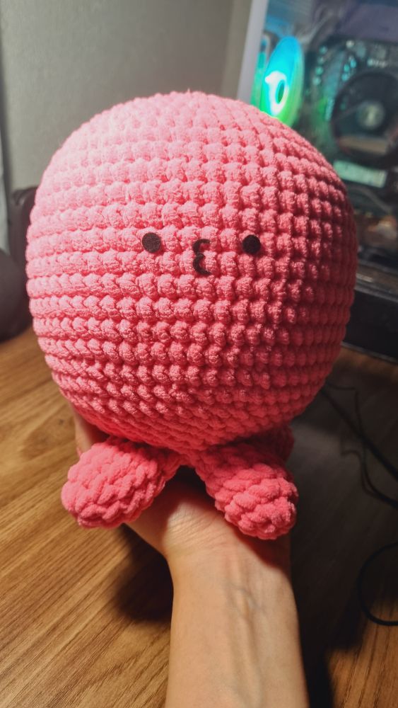Pelúcia do personagem Takopii, um polvo rosa com quatro tentáculos, ao fundo uma mesa com um gabinete em cima.