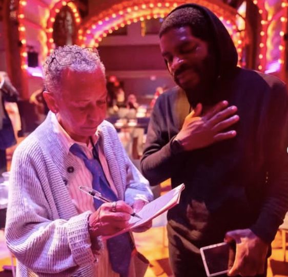 Photo of Nikki Giovanni and Hanif Abdurraqib. Per Hanif, "In 2023, after Giovanni performed at the Brooklyn Academy of Music, in a series I curated, somewhat selfishly, to my own tastes and desires, we took a few photos together. In the photo I love most, Giovanni is signing my copy of “Black Judgement,” which I’d nervously brought with me from Columbus to New York earlier that morning. She’s immersed in the process of signing her name." Hanif has his hand over his heart while Nikki signs.