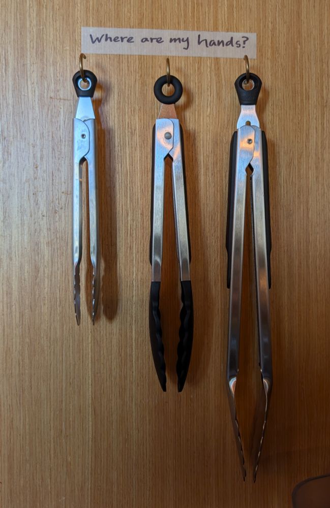 Three pairs of kitchen tongs hanging from hooks on a wooden panel. 7" metal tongs, 9" nylon tongs, and 12" metal tongs. Arranged smallest to largest from left to right. Above the tongs there is a strip of label tape with sharpie ink that says "Where are my hands?"