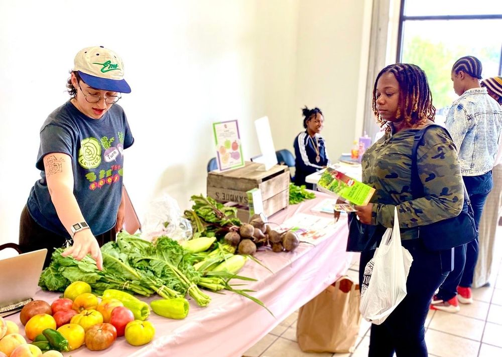 A vendor pointing at a table full of fruits and vegetables for a customer