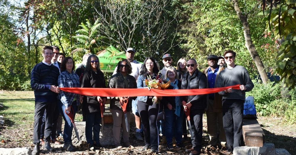 A team poses with a large red ribbon in front of green trees