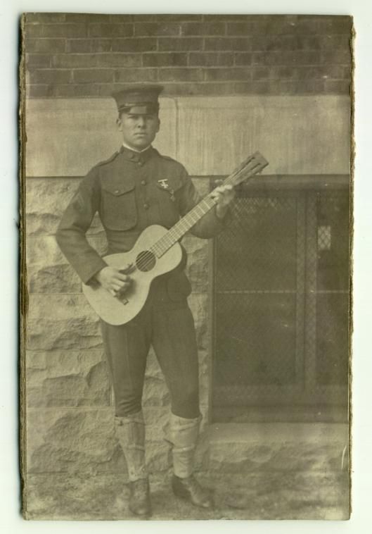 Soldier in uniform, c1900, standing in front a brick and stone building playing a guitar