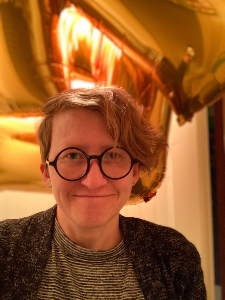 A woman wearing round glasses, a striped shirt, and a black and white speckled cardigan, smiles amid several gold Mylar balloons 
