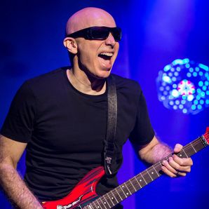 A bald man in wraparound sunglasses, black t-shirt plays a red electric guitar. He’s having a great time. 

He is Joe Satriani