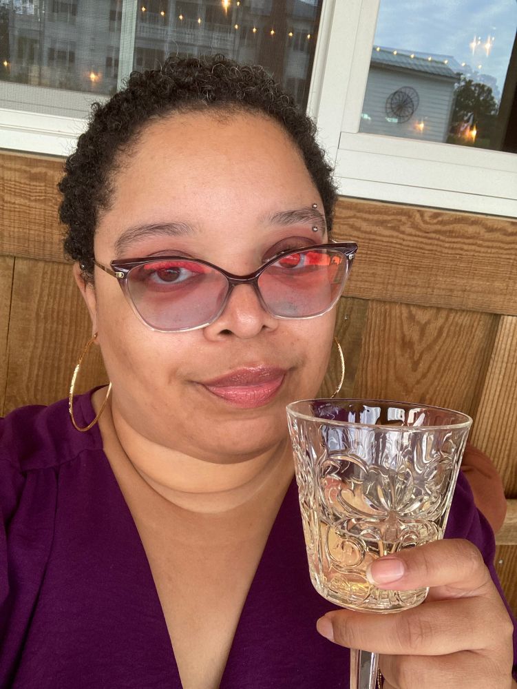 Indie stares at you through tinted glasses. She holds up a glass of champagne. She is wearing a plum dress. 