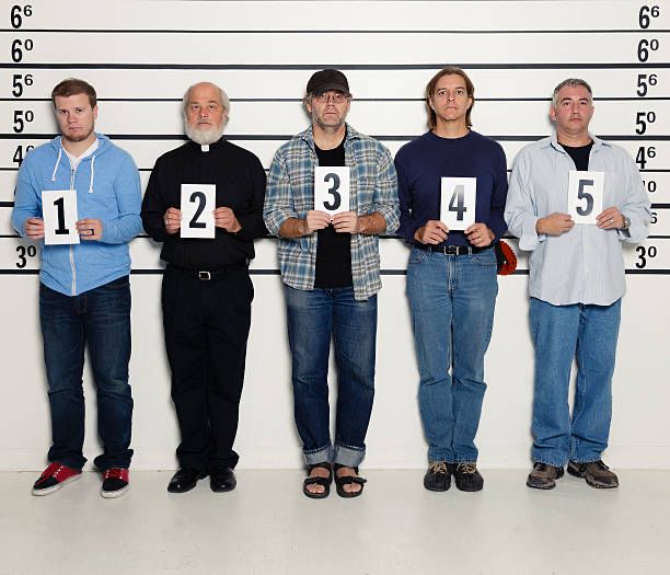 5 men stand in a police line up in front of a wall with height markings holding signs labeled 1 through 5. 