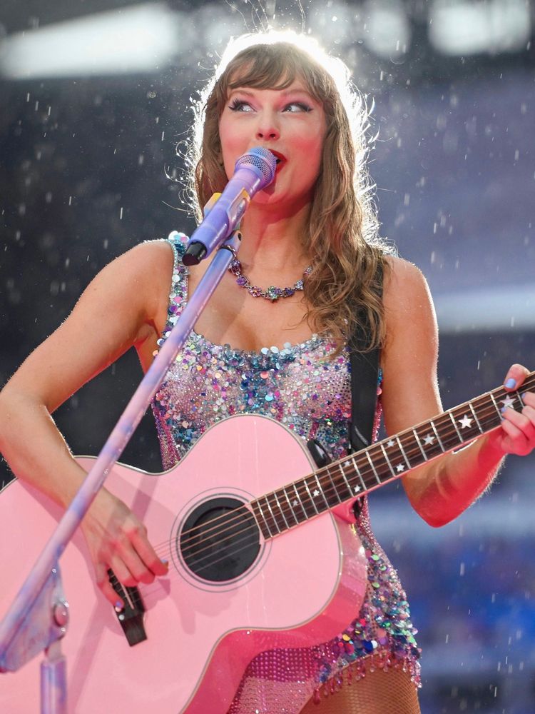 Taylor Swift on stage at the Eras Tour wearing the sparkling pink Lover bodysuit decorated with light blue sequins. It’s is raining and Taylor is looking playfully to the side, looking excited. She is holding a pink guitar.