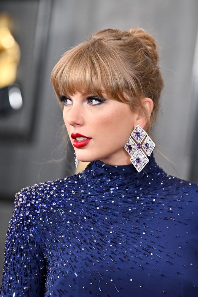 Taylor Swift photographed at the 2023 GRAMMYs red carpet, wearing a dark navy, long sleeved crop top and skirt, bejeweled with blue gems. She’s wearing her hair up, big dramatic earrings and red lipstick