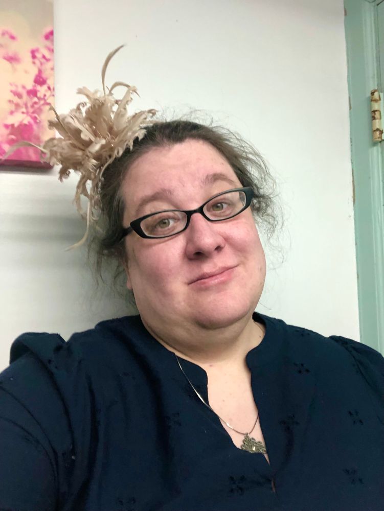 A selfie of me after a full day of funeral and funeral dinner. My hair is a little unkempt. I have on a navy blue dress and a champagne colored feather fascinator. My facial expression is a little melancholy and a lot tired.